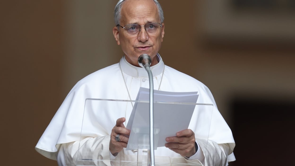 Pope Leo stands in front of a podium reading from a piece of paper. 